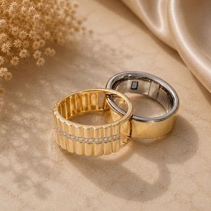 Peut inclure: Deux bagues entrelacées, l'une dorée avec une texture côtelée verticale et une bande centrale de petites pierres claires, et l'autre une bague dorée lisse et polie avec une doublure intérieure argentée et un petit capteur rectangulaire.