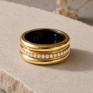Peut inclure: Une bague dorée dotée d'une bande centrale incrustée d'une rangée de petites pierres rondes transparentes. La bague présente une finition dorée lisse et polie sur les bords supérieur et inférieur, et l'intérieur est doublé de noir.