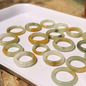 May include: A collection of smooth, circular rings crafted from natural stone, featuring a translucent pale green base with distinct patches of warm yellow and orange hues, arranged in a scattered pattern on a white surface.