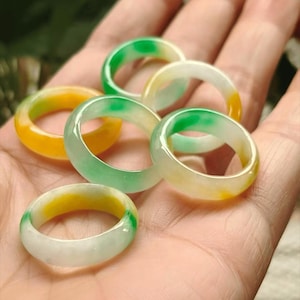 May include: A collection of five smooth, polished band rings featuring a mix of translucent white, vibrant green, and warm yellow hues. Each ring has a rounded, circular profile with a glossy finish, showcasing natural color variations.