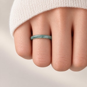 May include: A smooth, solid band ring crafted from polished green jade stone. The ring features a consistent, rounded profile with natural variations in the light and dark green mineral coloring throughout the surface of the circular band.