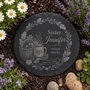 Puede incluir: Una piedra conmemorativa redonda de pizarra negra con un grabado de una cesta y una taza con la etiqueta CIDER. El texto dice Sister Jennifer Always Nearby April 28, 2022, rodeado por una corona de hojas de roble y bellotas en la superficie.