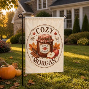 May include: A rectangular garden flag featuring a cream background with a central illustration of a glass jar of apple butter with a red and white checkered lid. The text reads COZY MORGAN. Surrounding the jar are cinnamon sticks and autumn leaves.