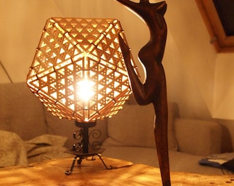 May include: A wooden sculpture of a woman in a dancing pose, with a wooden base, on a wooden table with a lamp with a geometric patterned shade.