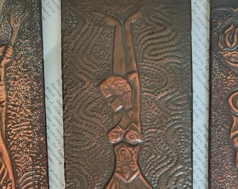 May include: Three copper wall plaques with embossed designs. The centre plaque features a woman in a ballet pose. The plaque on the left has a tree design. The plaque on the right has a church and water design.