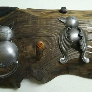 May include: A wooden wall-mounted coat rack with three metal hooks. The hooks are shaped like a cat, a bird, and a simple hook. The wood is dark brown with a natural finish.