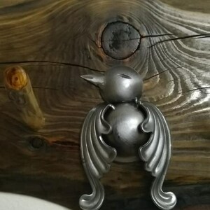 May include: A silver metal bird-shaped hook with wings and a round body, mounted on a rustic wooden surface.