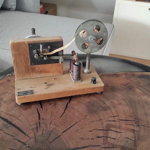 May include: A vintage wooden device with a metal spool and a crank. The device is used for winding wire or thread. It is sitting on a wooden table with a dark brown finish.