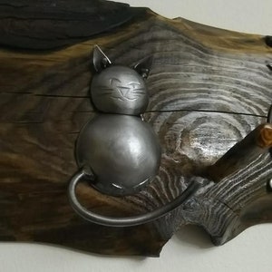 May include: A metal cat figurine with a smiling face, attached to a wooden wall hanging. The cat is made of metal and has a silver finish. The wooden wall hanging is brown and has a natural wood grain.