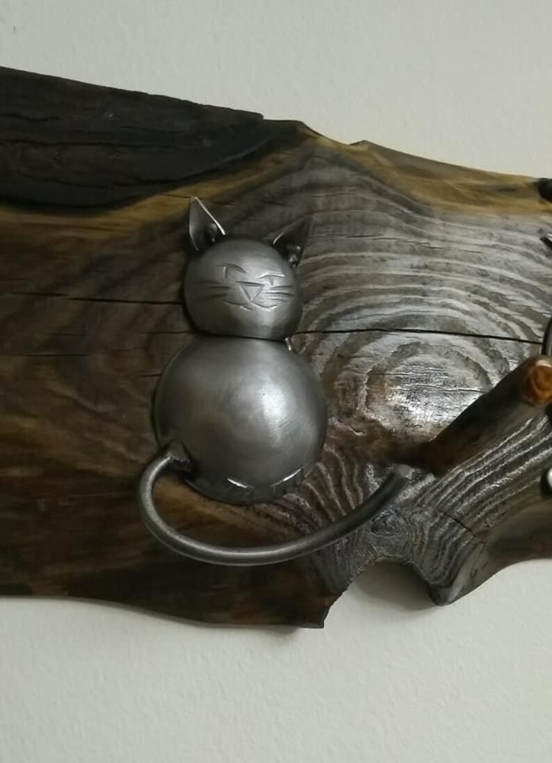 May include: A metal cat figurine with a smiling face, attached to a wooden wall hanging. The cat is made of metal and has a silver finish. The wooden wall hanging is brown and has a natural wood grain.
