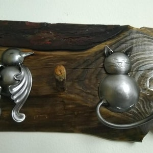 May include: A rustic wooden wall hook with a metal bird and cat design. The bird is made from a spoon and has a long, curved beak. The cat is made from a spoon and has a round body and a long, curved tail.