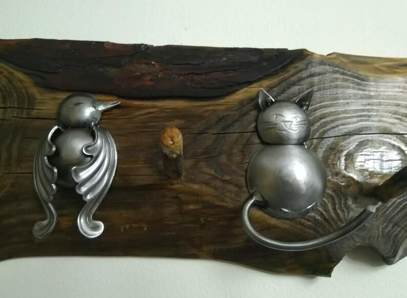 May include: A rustic wooden wall hook with a metal bird and cat design. The bird is made from a spoon and has a long, curved beak. The cat is made from a spoon and has a round body and a long, curved tail.