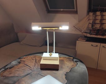 Könnte beinhalten: Eine weiß-goldene Schreibtischlampe mit rechteckigem Sockel und doppeltem Leuchtenkopf. Die Lampe steht auf einem Holztisch mit einzigartigem, organischem Design. Ein Modellsegelschiff ist im Hintergrund zu sehen.