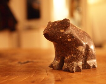 May include: A small, brown, speckled stone sculpture of a bear sitting on a wooden surface.