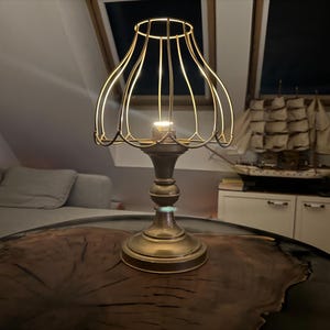 Può includere: Una lampada da tavolo in metallo dorato con un paralume a gabbia di filo. La lampada è su un tavolo di legno con una finitura marrone scuro.