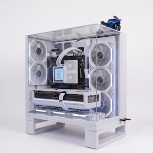May include: A white desktop computer tower with a glass front and side panel, featuring internal white cooling fans, a liquid cooling system, and a digital display showing 61 degrees. A small blue motorcycle model sits on top of the computer case.