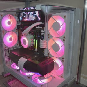 Puede incluir: Una torre de ordenador blanca con panel lateral de cristal, con componentes internos iluminados por ventiladores LED rosas y blancos. El sistema incluye un bloque de refrigeración líquida, cables trenzados blancos y una tarjeta gráfica negra.
