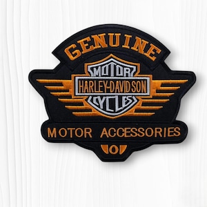 May include: A black embroidered patch featuring the text GENUINE in orange, a central shield logo with the text MOTOR HARLEY-DAVIDSON CYCLES in white and orange, and the text MOTOR ACCESSORIES in orange at the bottom of the design.
