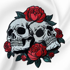 May include: An embroidered patch featuring two white skulls with black eye sockets and teeth, surrounded by vibrant red roses and dark green leaves. The design is detailed with black stitching outlines, creating a high-contrast graphic element.