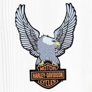 May include: An embroidered patch featuring a bald eagle with wings spread wide, perched atop a black and orange shield-shaped emblem. The emblem displays the text MOTOR HARLEY-DAVIDSON CYCLES in orange lettering against a black background.
