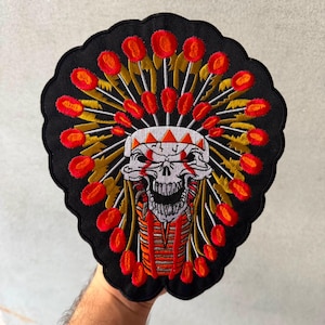 May include: A black embroidered patch featuring a central white skull with red accents, surrounded by a radiating sunburst pattern of gold rays and vibrant red oval shapes. The patch has a scalloped edge and a detailed, multi-colored thread design.