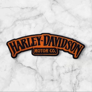 May include: An arched embroidered patch featuring the text Harley-Davidson in black lettering on an orange background, with the words Motor Co. centered below in a smaller black font, all outlined with a thick black border.