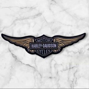 May include: An embroidered patch featuring the Harley-Davidson logo with a central shield and extended wings. The design uses black, white, and gold thread, with the text MOTOR HARLEY-DAVIDSON CYCLES clearly visible in the center.
