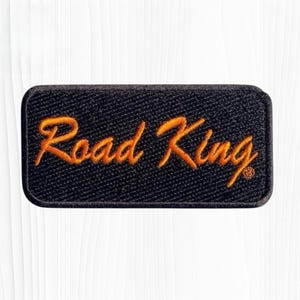 May include: A rectangular embroidered patch with a black background and a dark border. The text Road King is stitched in a bright orange cursive font across the center of the patch. The surface has a visible fabric texture throughout.
