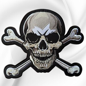 May include: An embroidered patch featuring a stylized human skull centered over two crossed bones. The design uses shades of beige, white, and black thread with a thick black border outlining the entire shape against a neutral background.