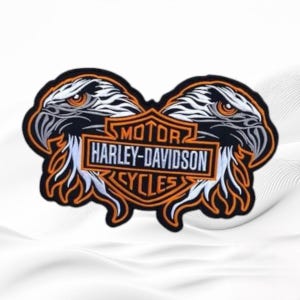 May include: An embroidered patch featuring two eagle heads facing outward, flanking a central orange and black shield emblem with the text MOTOR HARLEY-DAVIDSON CYCLES. The design uses black, white, and orange thread on a white fabric background.