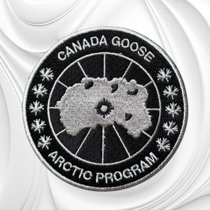 May include: A circular embroidered patch featuring a black background with a silver-grey map of the Arctic region in the center. The text CANADA GOOSE and ARCTIC PROGRAM are stitched in white along the border, accented by small maple leaf icons.