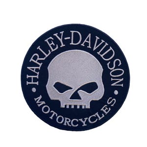 May include: A circular embroidered patch featuring a dark blue border with the white text Harley-Davidson Motorcycles. The center of the patch displays a light gray skull graphic with dark blue eye sockets and teeth details.