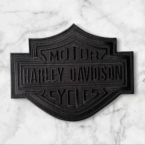 May include: A black embroidered patch in the shape of the Harley-Davidson shield logo. The patch features the text MOTOR HARLEY-DAVIDSON CYCLES stitched in black thread, creating a monochromatic design with raised lettering and a defined border.