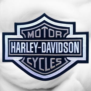 May include: A shield-shaped embroidered patch featuring the text MOTOR HARLEY-DAVIDSON CYCLES in white and light gray thread against a black background. The design includes a thick white border and a distinct bar-and-shield emblem shape.
