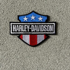 May include: A shield-shaped embroidered patch featuring the text HARLEY-DAVIDSON in white letters on a black rectangular background. The top section is blue with three white stars, and the bottom section has vertical red and white stripes.