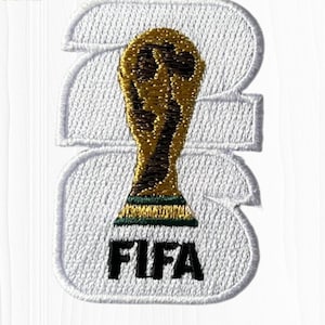 May include: A white embroidered patch featuring the number 2026 in a bold font. Centered over the numbers is a gold and green FIFA World Cup trophy icon. The word FIFA is stitched in black thread at the bottom of the white embroidered background.