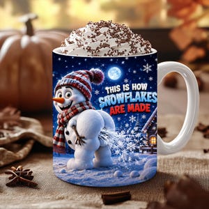 Puede incluir: Una taza de cerámica blanca llena de nata montada y virutas de chocolate. La taza presenta un gráfico de un muñeco de nieve con bufanda y gorro de cuadros rojos y blancos, con el texto "THIS IS HOW SNOWFLAKES ARE MADE" en azul y rojo.