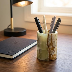Peut inclure: Un porte-stylo cylindrique en pierre présentant une surface polie avec des motifs marbrés dans des tons de vert clair, crème et brun foncé. Le support contient quatre stylos de couleur noire, blanc cassé, gris clair et gris foncé, placés verticalement.