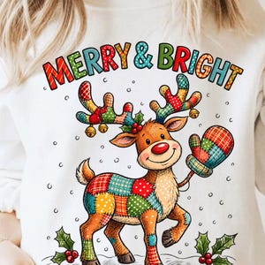 Può includere: Una felpa bianca con una grafica colorata di una renna con corpo e corna in stile patchwork. La renna indossa una muffola abbinata e ha il naso rosso. Il testo MERRY & BRIGHT è stampato sopra in lettere multicolori.