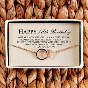 Peut inclure: Un collier en or rose avec deux cercles imbriqués est présenté dans une boîte blanche. La boîte contient une carte avec le texte "HAPPY 18th Birthday" et un message. Le fond est un motif tissé et texturé dans des tons bruns.