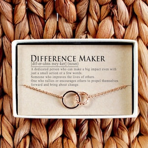 May include: A gold-colored necklace with two interlocking circles, presented in a white box against a woven background. The box features the text "DIFFERENCE MAKER" and its definition.