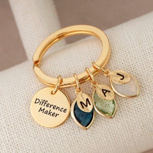 May include: A gold-coloured keyring with a round charm inscribed with "Difference Maker." The keyring also features three leaf-shaped charms with the letters "M," "A," and "J." The leaf charms are in shades of blue, green, and white.