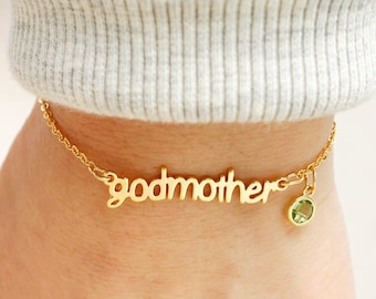 May include: A gold-toned chain bracelet featuring the word godmother in a cursive script font. The bracelet includes a small, round, light green faceted gemstone charm attached to the chain next to the lettering, creating a delicate accessory.