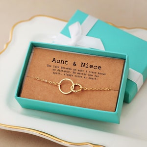 May include: A turquoise gift box with a white ribbon contains a gold bracelet with two intertwined circles. The box has a card inside with the text "Aunt & Niece. The love between an aunt & niece knows no distance. No matter how far apart, always close at heart."