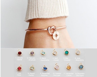 May include: A rose gold bangle bracelet with a knot design and a compass charm. The compass charm has a blue star in the center. The bracelet is shown with 12 different charms, each representing a different month of the year and birthstone. The charms are shown in a row below the bracelet.