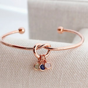 May include: A rose gold bangle bracelet with a knot design and a charm with three small stones. The stones are green, blue, and pink.