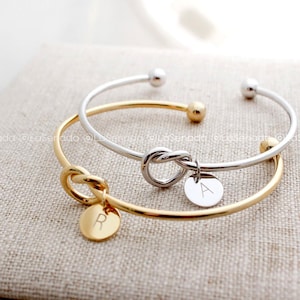 May include: Two gold and silver knot bracelets with initial charms. The gold bracelet has a charm with the letter "R" and the silver bracelet has a charm with the letter "A".