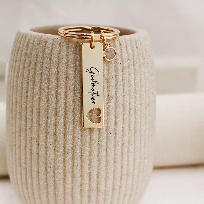 May include: Gold-toned keychain with a rectangular tag that says "Godmother" with a heart cut-out. The keychain also has a small clear crystal charm.