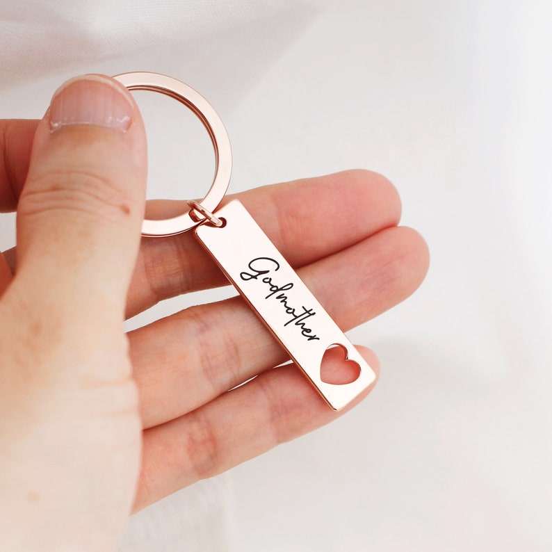 May include: Rose gold keychain with a heart cut-out and the word "Godmother" engraved on it.