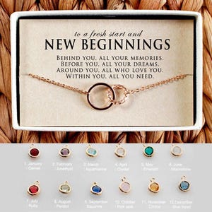 Könnte beinhalten: Eine goldene Halskette mit zwei ineinander verschlungenen Kreisen, ein Symbol für Neuanfänge. Die Halskette wird in einer weißen Schachtel mit einer Botschaft über Neuanfänge präsentiert. Unter der Halskette befinden sich 12 kleine, farbige Edelsteine, die die Monate des Jahres repräsentieren.